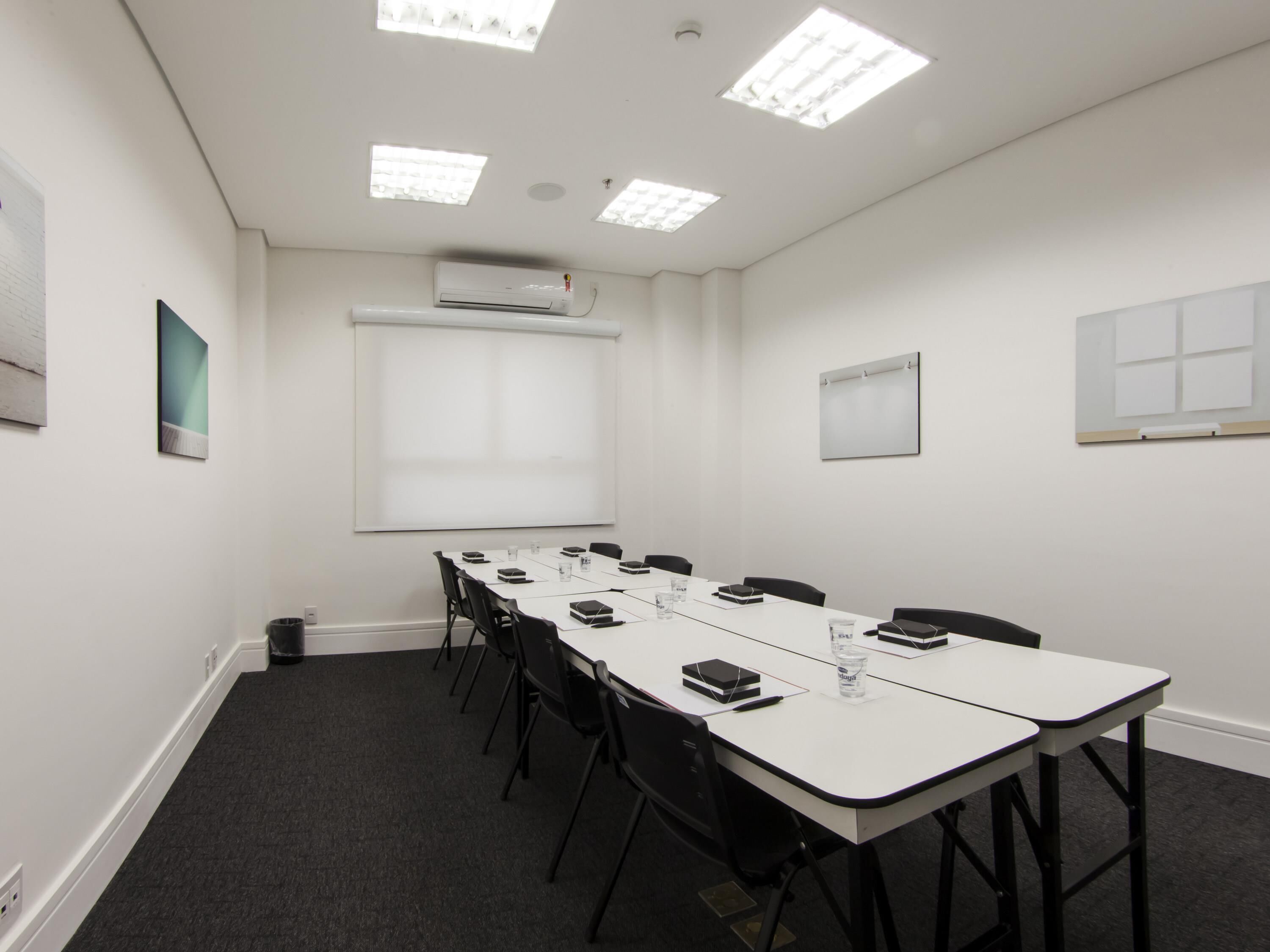 Una sala de conferencias con una larga mesa, sillas negras, una pantalla de proyector y una pizarra.