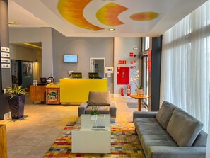 Een hotel lobby met een receptiebalie, een zithoek, een brandblusser en een televisiescherm.