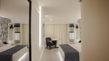 Habitación de hotel moderna con un armario espejado, cama con cubrecama estampado, pequeño escritorio con silla y mesita de noche con lámpara.