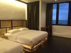 Ein Hotelzimmer mit zwei Betten auf Holzböckchen, einem kleinen Tisch und Meerblick durch große Fenster.