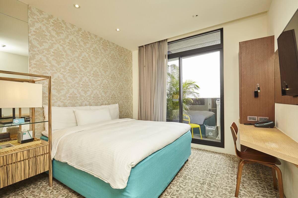 A hotel room with a bed, a desk, a chair, a mirror, and a window with a view of palm trees.