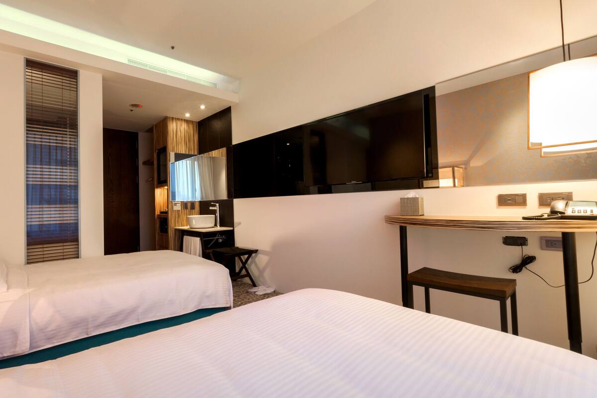 A hotel room with two beds, a TV, a desk with a lamp, and a bathroom with a sink and mirror.