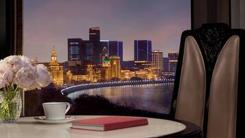 A table with a cup, saucer, book, and vase of flowers, with a cityscape view through a window.