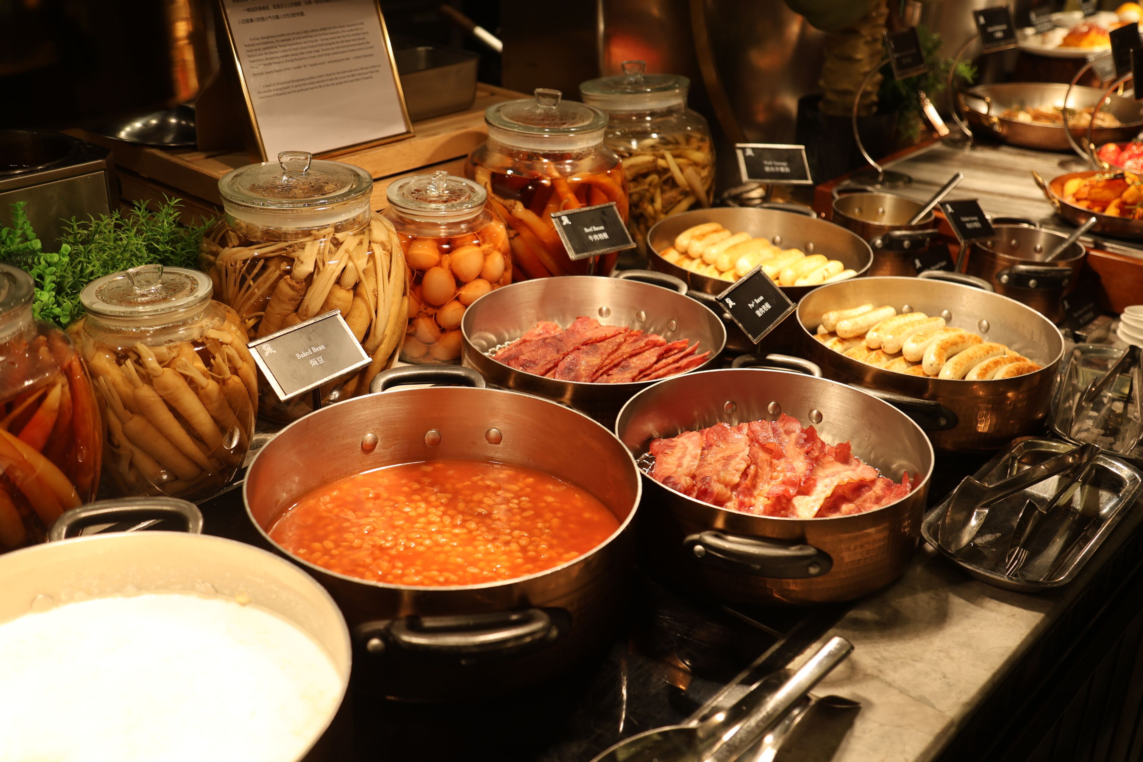 En buffet med forskellige retter i metalbeholdere, blandt andet bønner, kød og grøntsager.