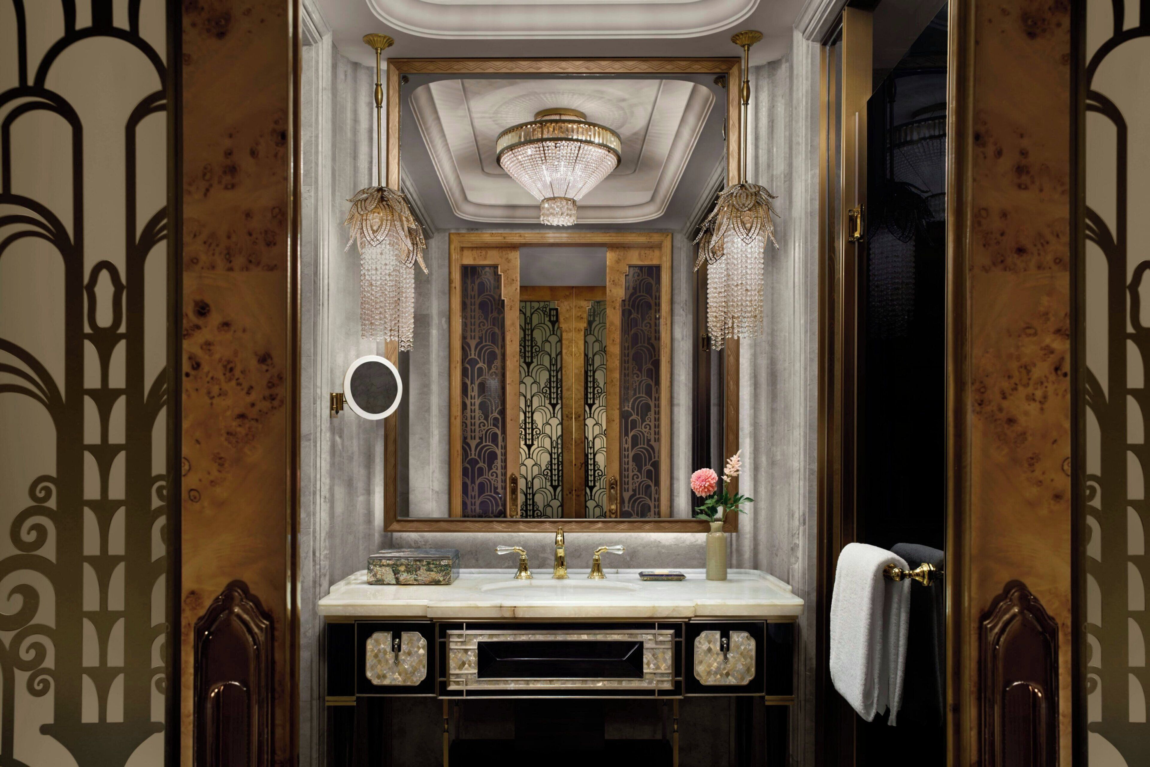 A luxurious bathroom with a large mirror, ornate wall panels, a chandelier, and a vanity with a gold faucet.