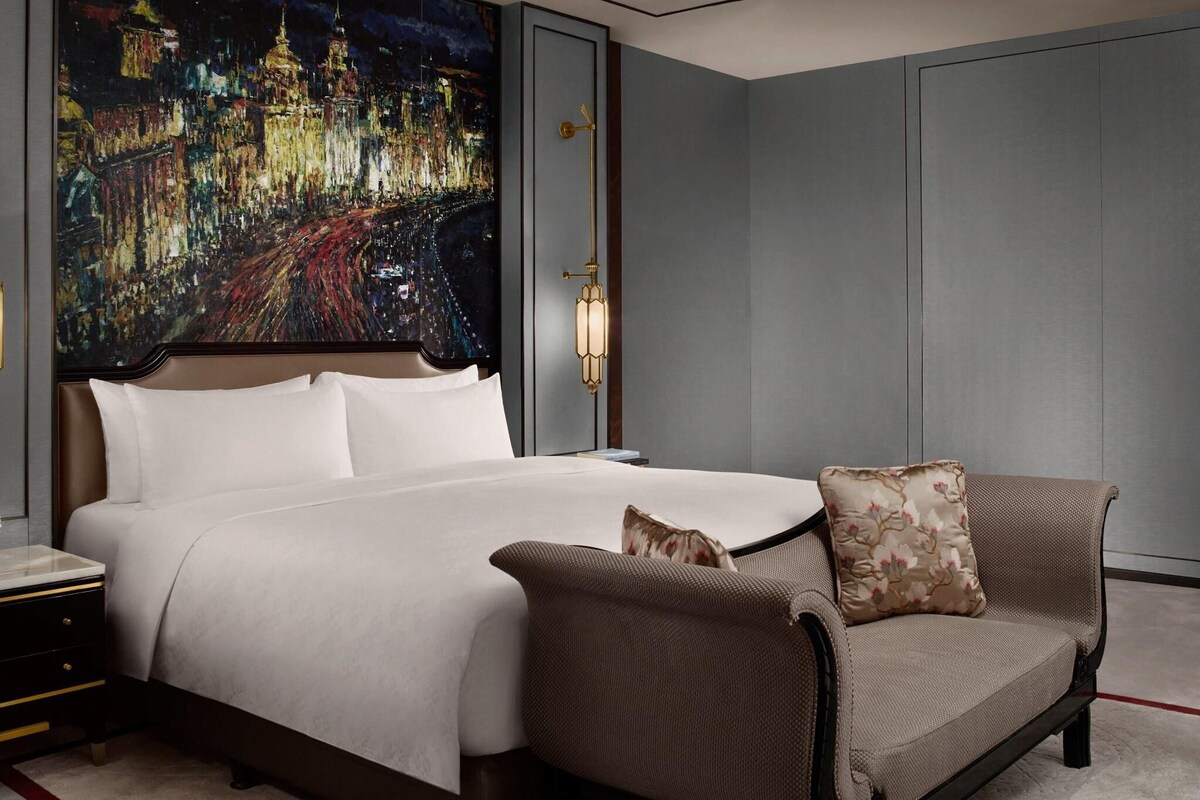 A hotel room with a large bed, a night cityscape painting on the wall, a nightstand, and a sofa with a cushion.