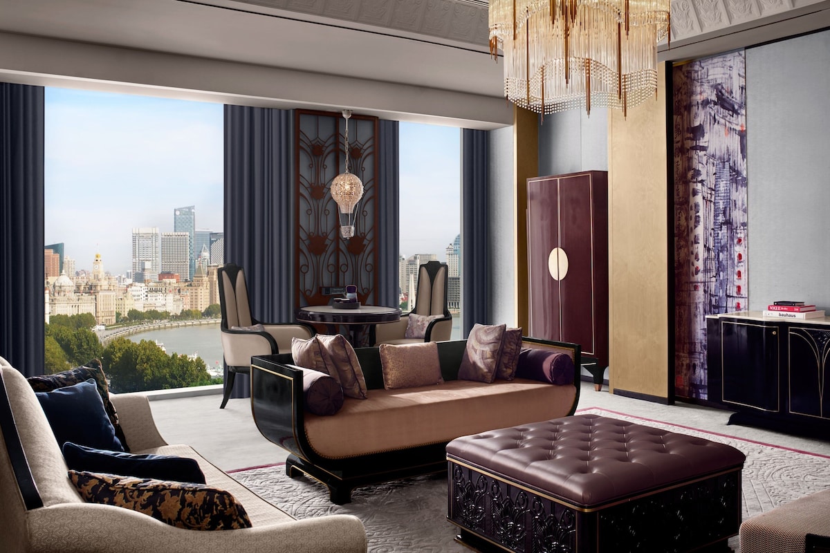 A modern living room with a sofa, ottoman, and a dining area with a table and chairs. A large window offers a city view.