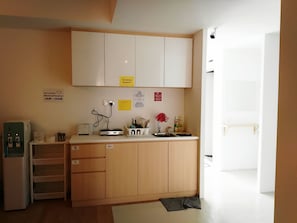 Una pequeña cocina equipada con refrigerador, cafetera y lavamanos. Hay carteles que indican los horarios del desayuno y las instrucciones de limpieza.