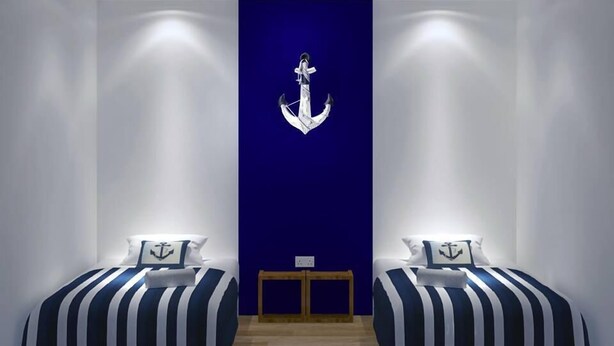 Habitación con dos camas, cada una con ropa de cama a rayas y almohadas con forma de ancla, un banco de madera y una pared azul con una ancla blanca.