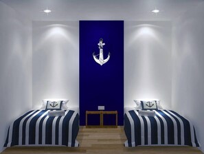 A room with two beds, each with striped bedding and anchor pillows, a wooden bench, and a blue accent wall with a white anchor. - Roof Talk Theme Hostel (Johor Bahru)