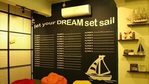 Una habitación con una pared grande que muestra una lista de sueños e una ilustración de un velero, puffs en rojo, verde y amarillo, y un estante pequeño con objetos decorativos.