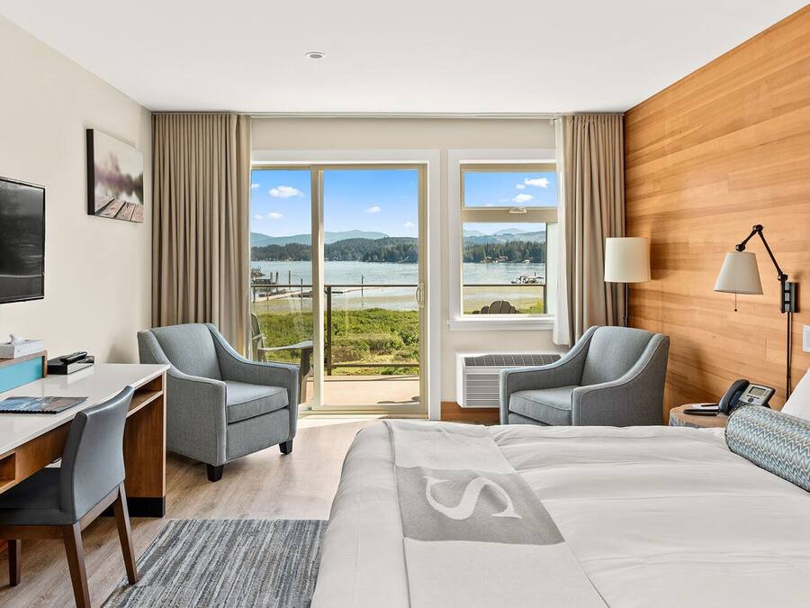 A hotel room with a bed, two armchairs, a desk, a TV, and a view of a lake and mountains through large windows.