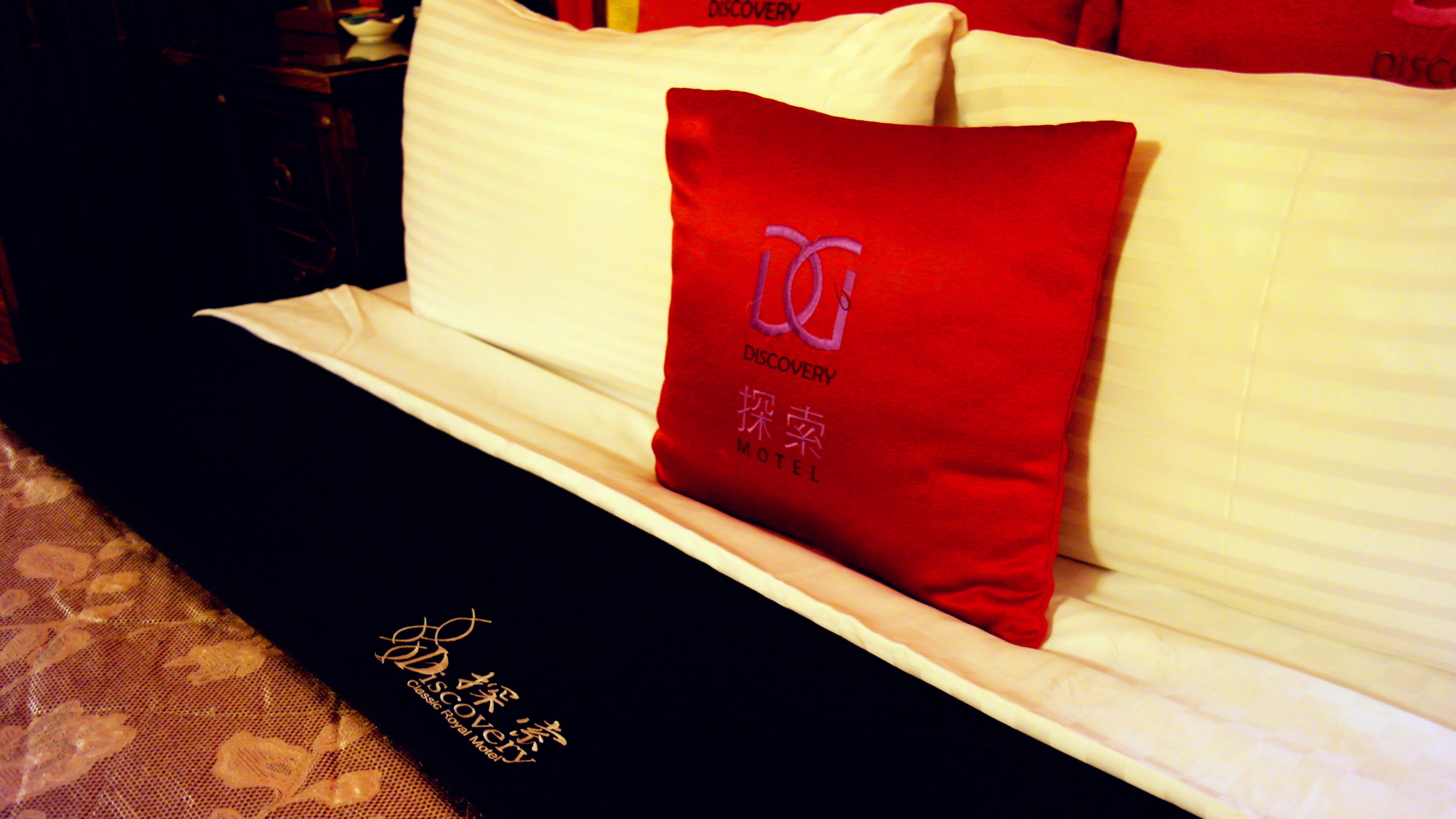 A bed with a white mattress and a red pillow with embroidered text.