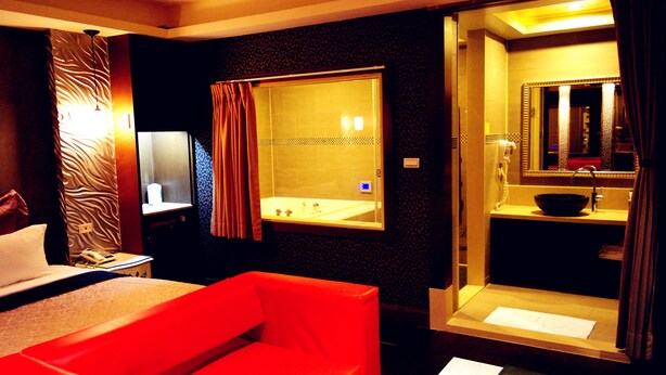 A hotel room with a bed, a red chair, a bathroom with a sink and mirror, and a shower area.
