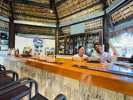 A bar area with a wooden counter, bar stools, and a thatched roof ceiling.