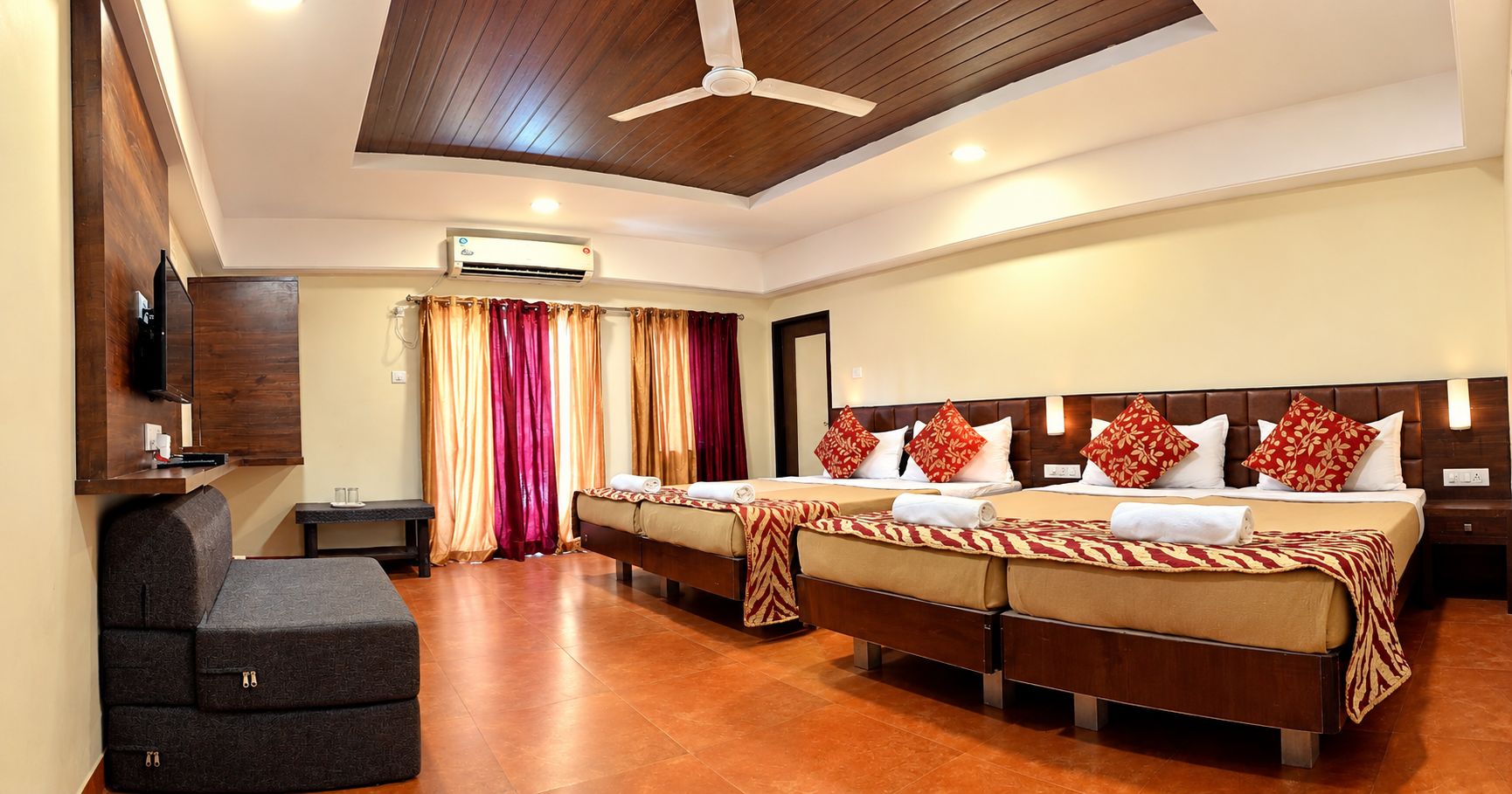 A hotel room with two beds, a sofa, a flat-screen TV, an air conditioning unit, and a ceiling fan.