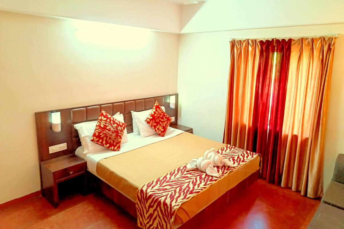 Una cama bien hecha con una manta de estampado de cebra y dos almohadas rojas, una mesita de noche con una lámpara, y una ventana con cortinas.