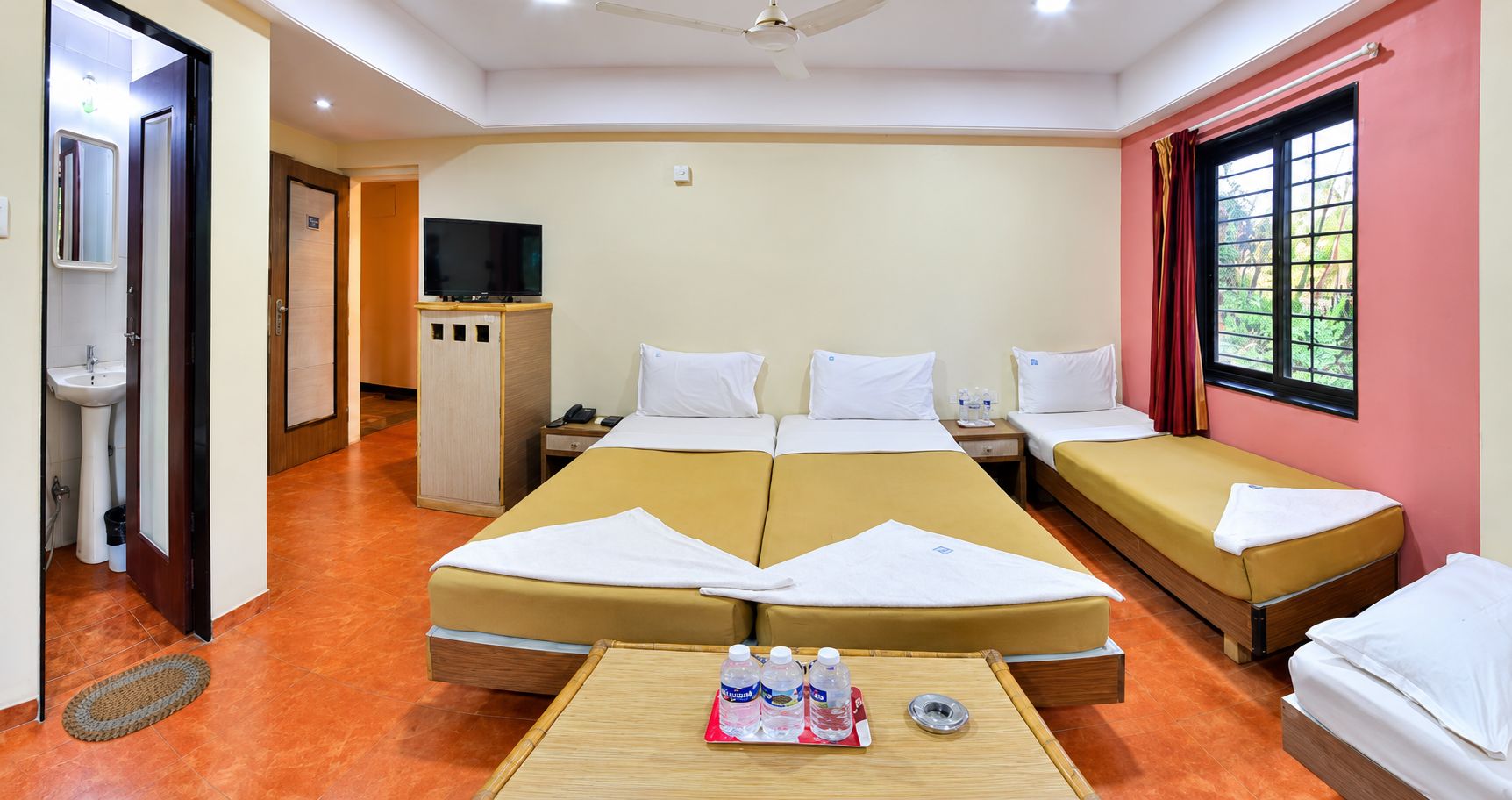 A hotel room with a large bed, two single beds, a TV, a small desk, and a window with curtains.