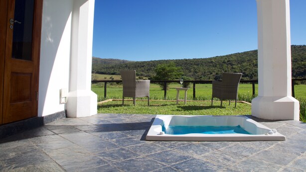Uma área externa coberta com uma banheira de hidromassagem, duas cadeiras de vime e vista para uma paisagem verde e colinas.
