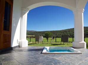 Un espace extérieur abrité comprenant un jacuzzi, deux fauteuils en osier et une vue sur un paysage verdoyant et des collines.