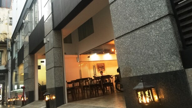 Un edificio con fachada de piedra, un área de estar al aire libre cubierta con mesas y sillas, y una entrada visible.