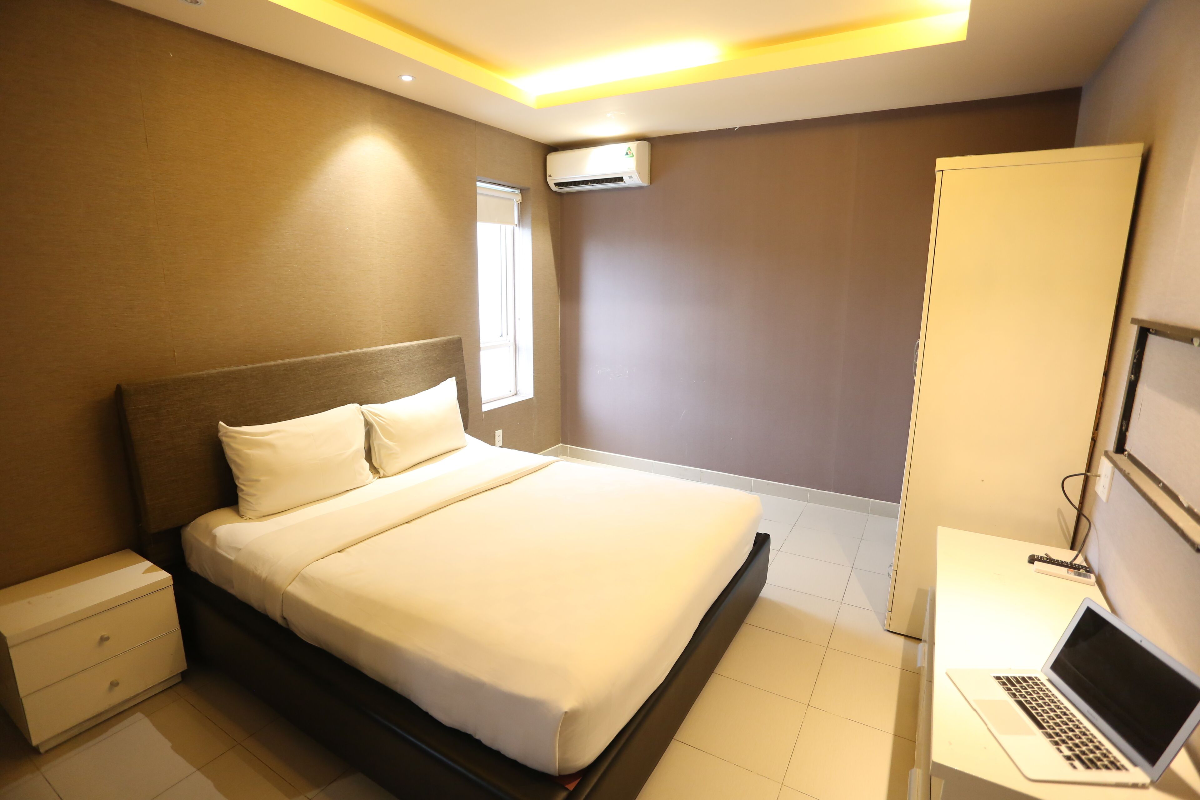 A modern hotel room with a neatly made bed, a bedside table with a laptop, and an air conditioning unit on the wall.