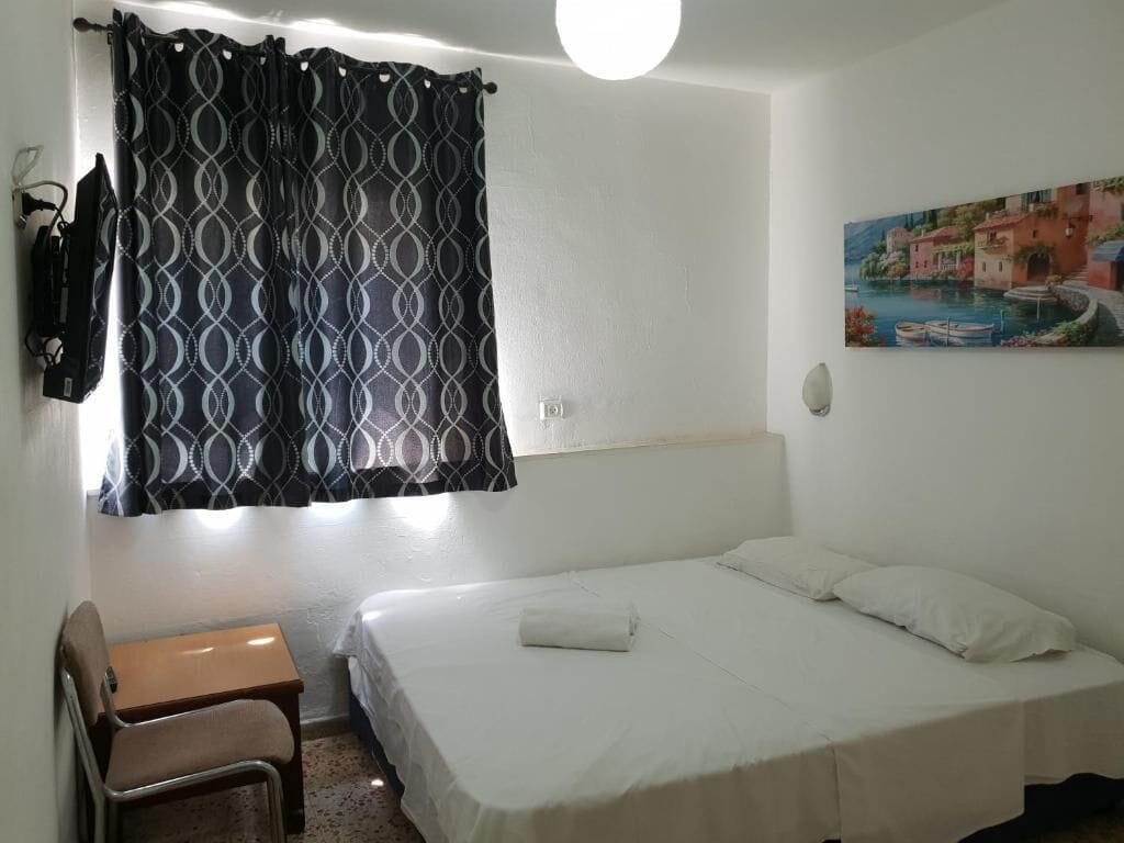 Una cama individual con colcha blanca, una mesita de madera, una silla, una cortina estampada y un cuadro con una escena costera en la pared.