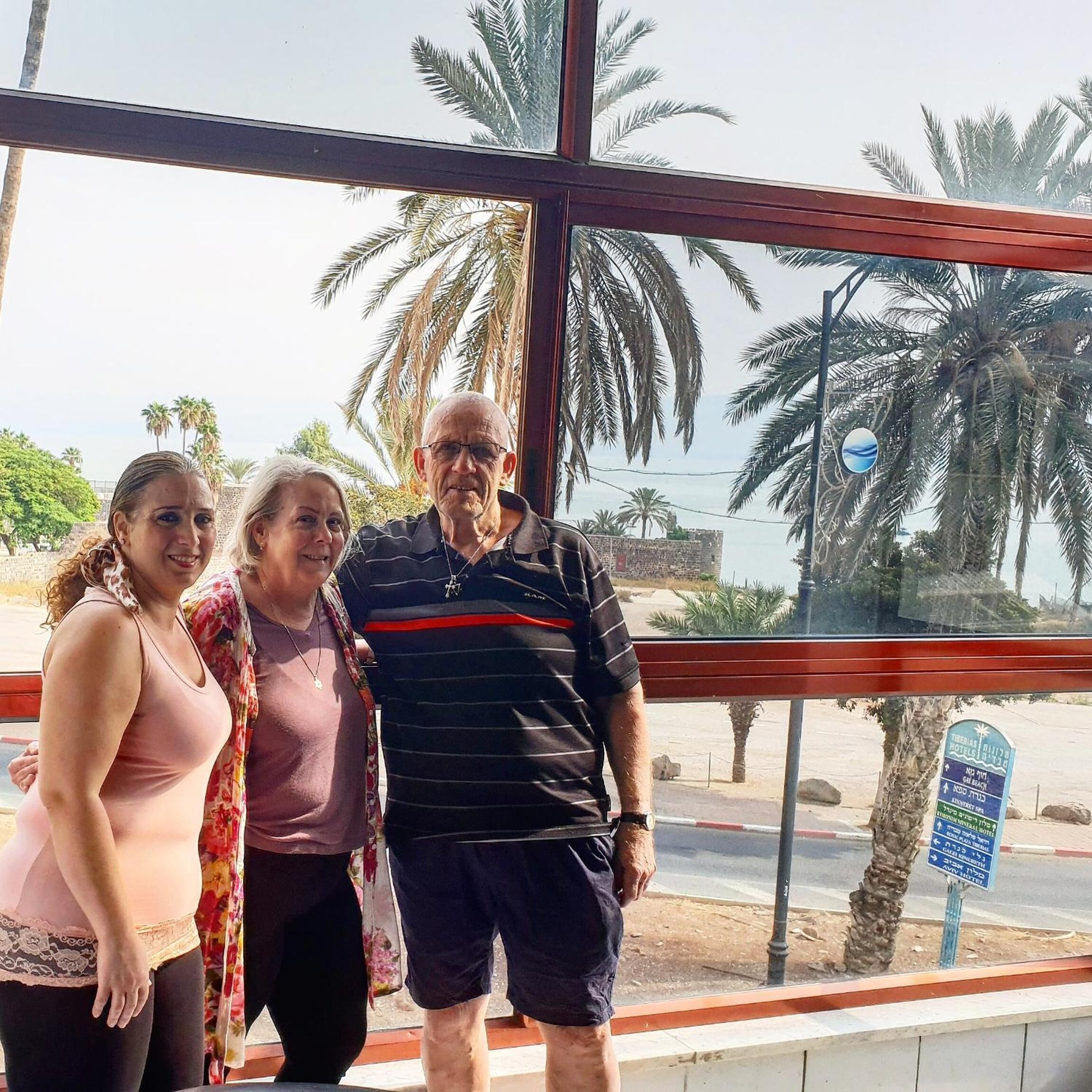Un grupo de personas de pie frente a un amplio ventanal con palmeras visibles al exterior.