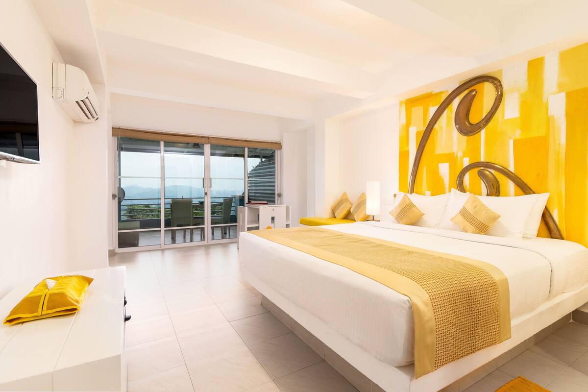 A modern hotel room with a large bed, a balcony with a view, and a bright yellow accent wall.