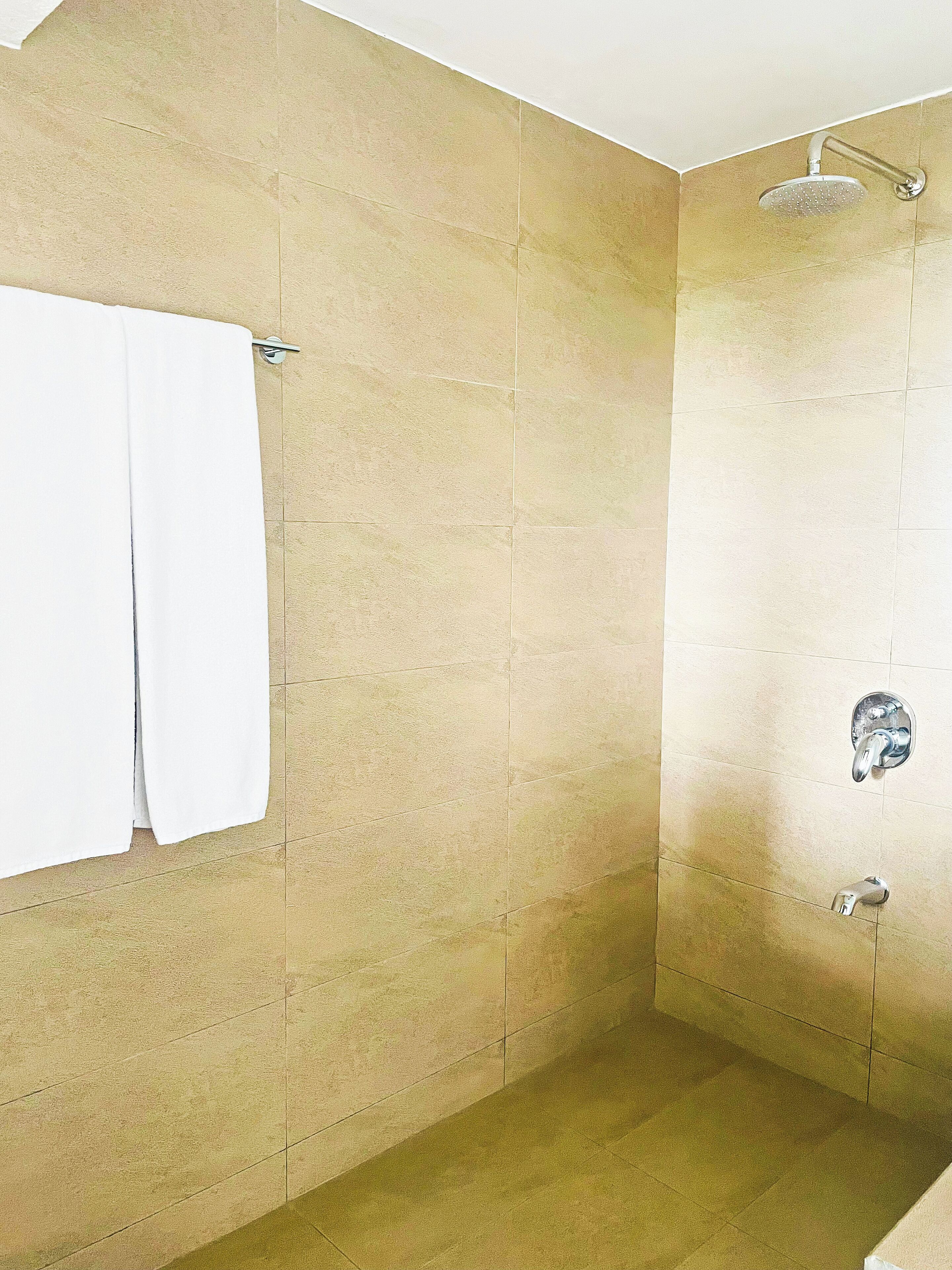 Un baño con baldosas color crema, una toalla blanca colgada en un tendedero y una ducha con cabezal de ducha manual.