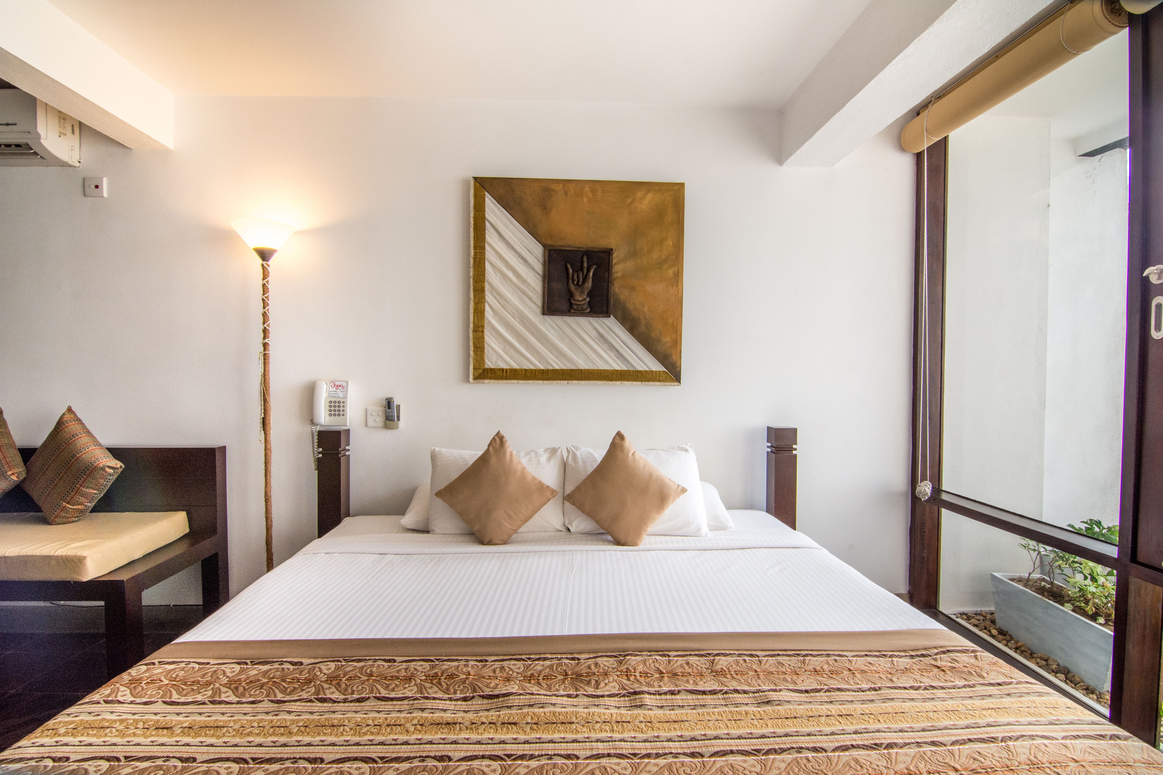 Habitación de hotel con una cama grande, una mesita de noche, una lámpara, teléfono en la pared y una ventana con vistas a zonas verdes.