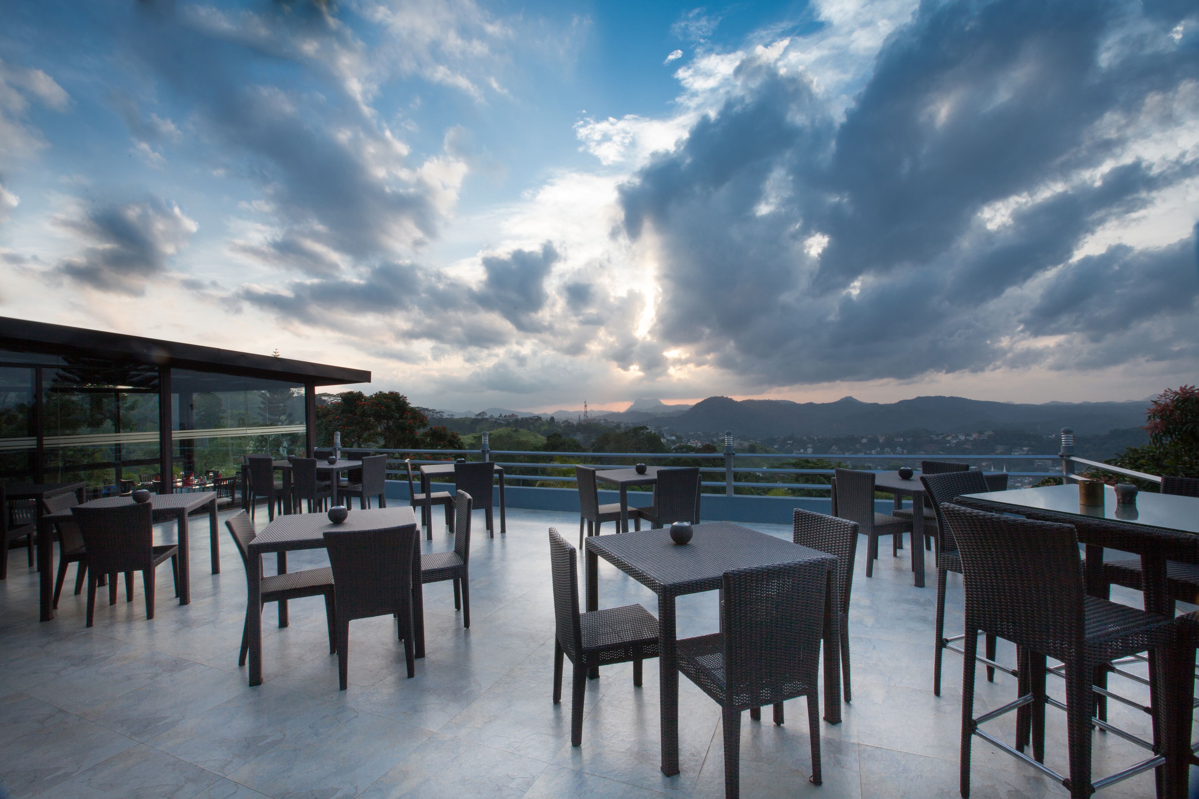 An outdoor terrace with tables and chairs, overlooking a scenic landscape with mountains and a body of water.