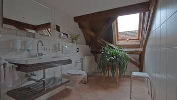 A modern bathroom with a large mirror, a sink, a toilet, and a shower area. There is a window providing natural light and a potted plant.