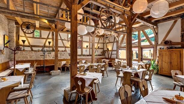 A rustic dining area with wooden tables and chairs, stone flooring, and exposed wooden beams. - Landhaus Zum Falken (Adelshofen)