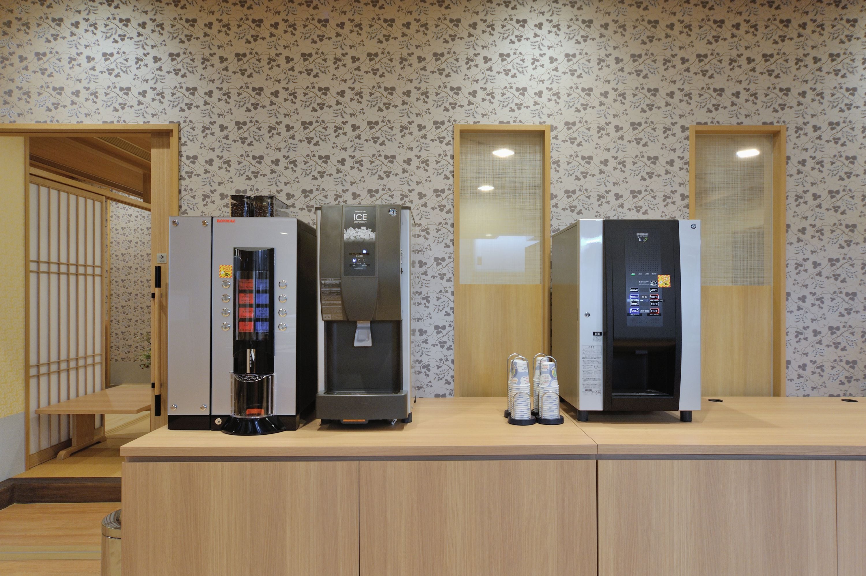 Una barra con tres dispensadores de bebidas y una máquina de café, frente a una pared con estampado floral.