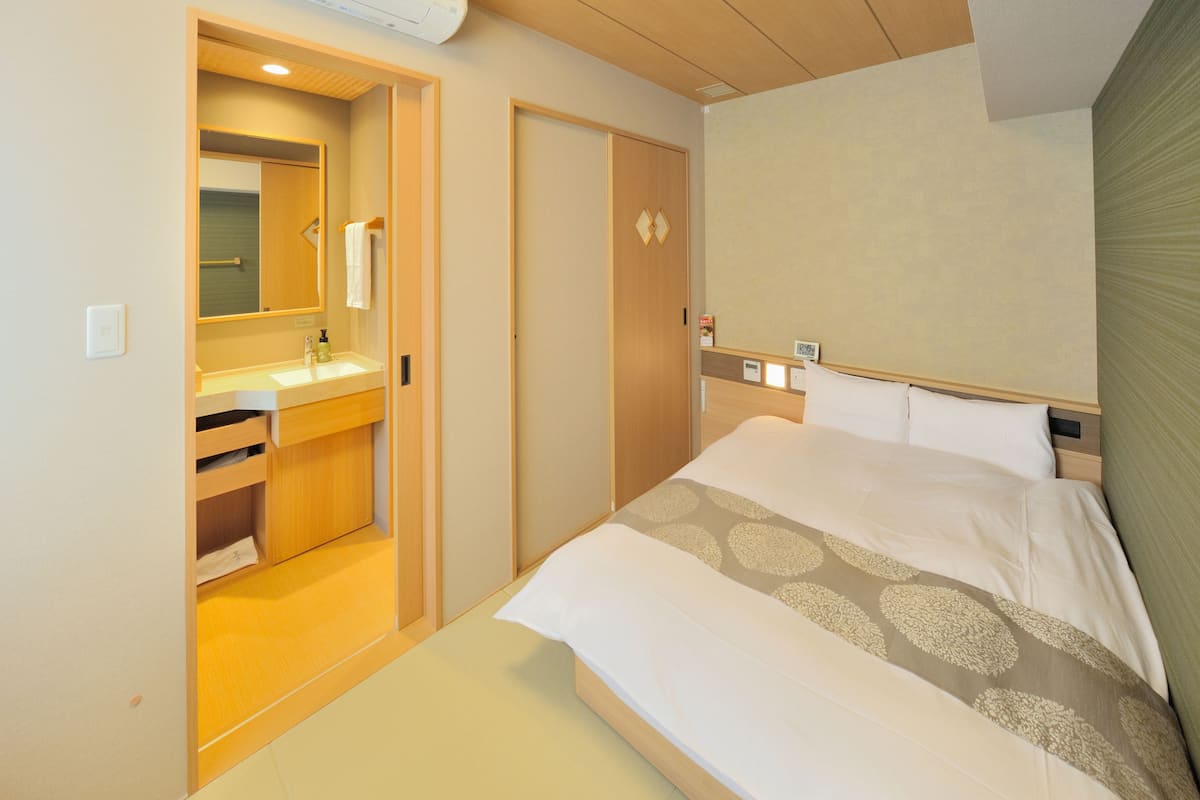 Habitación de hotel con cama, armario de madera, baño con espejo y una pequeña estantería.