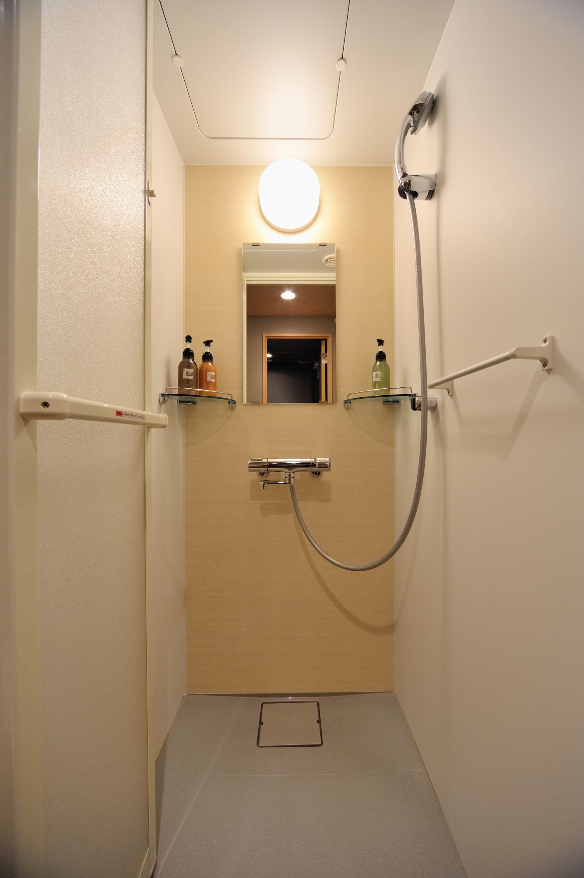 A narrow bathroom with a shower, a toilet, and a small mirror. There are two bottles of soap on a shelf.