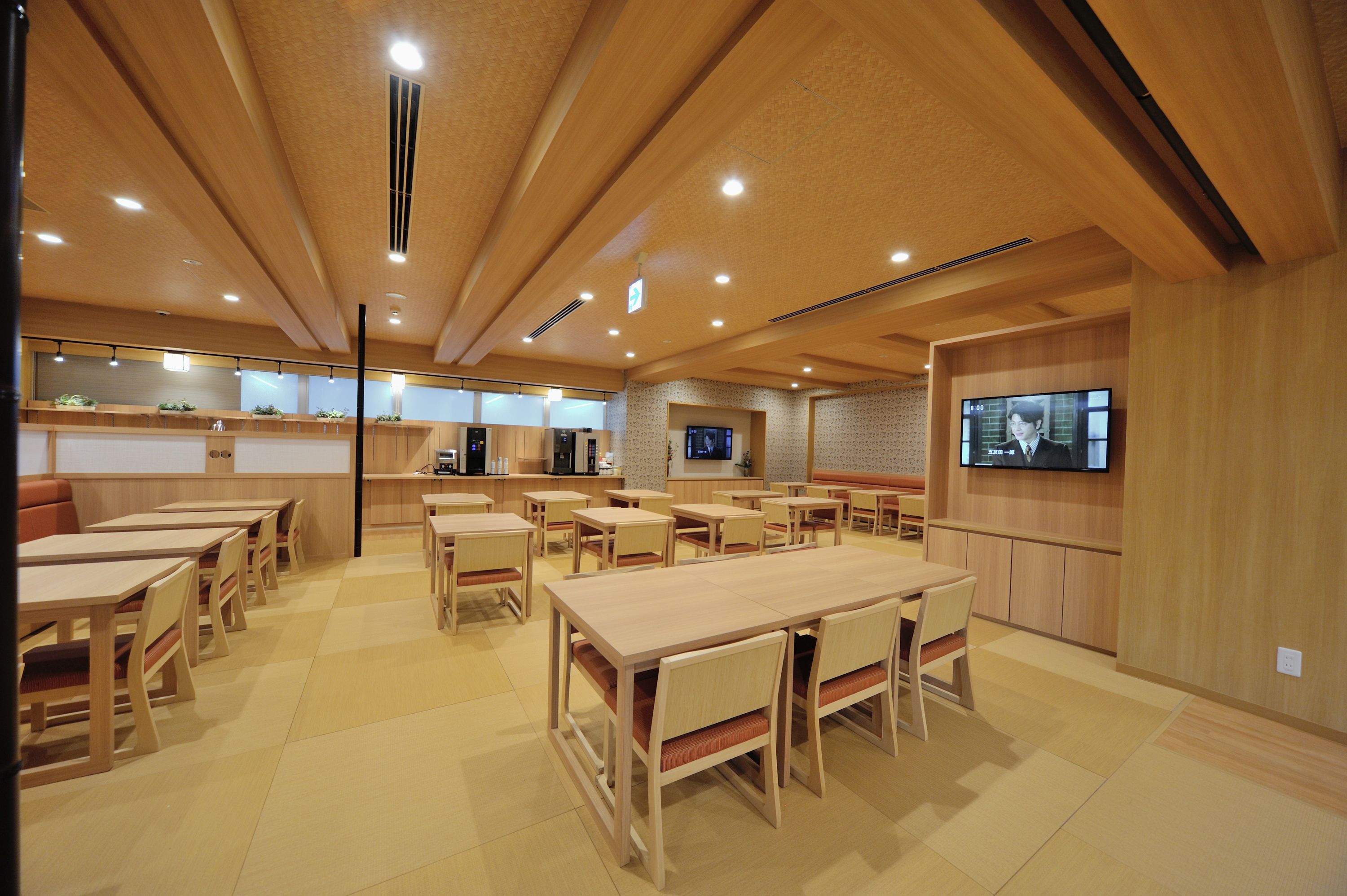 A spacious dining area with multiple wooden tables and chairs, a television mounted on the wall, and a kitchen area in the background.