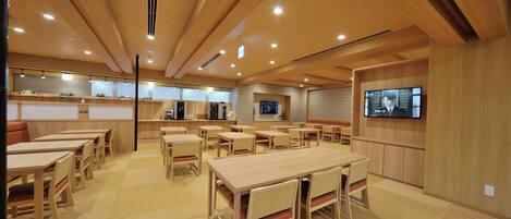 A spacious dining area with multiple wooden tables and chairs, a television mounted on the wall, and a kitchen area in the background.