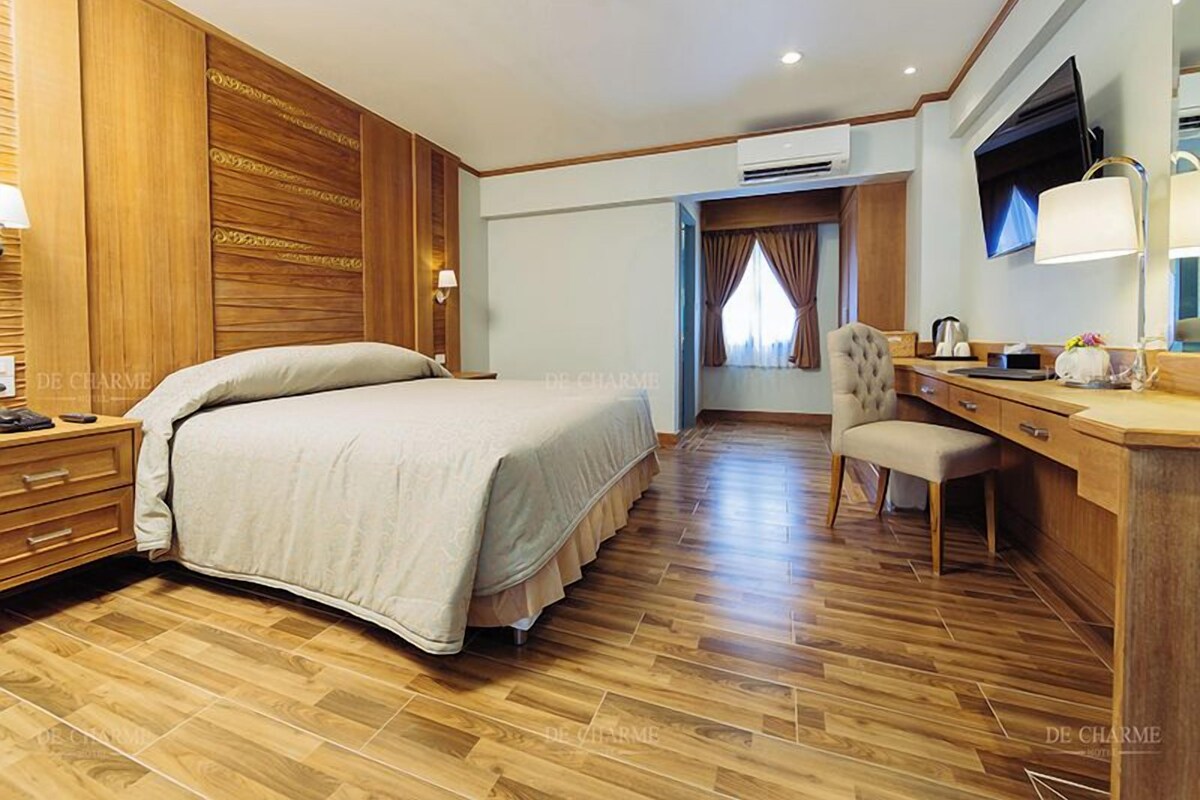 A hotel room with a wooden floor, a bed with a beige bedspread, a desk with a chair, a flat-screen TV, and a window with curtains.