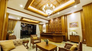 Un amplio vestíbulo de hotel con techo alto, paneles de madera ornamentados, un candelabro y una recepción.