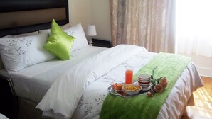 Un lit bien fait, avec du linge de lit blanc et des oreillers verts, une table de chevet avec une lampe et un plateau contenant des éléments pour le petit-déjeuner.