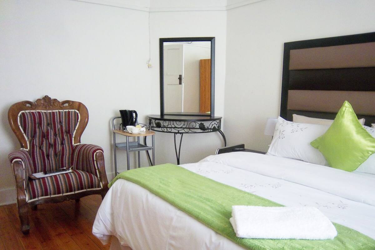 Habitación de hotel con cama, una silla, una mesita de noche con espejo y una manta verde.