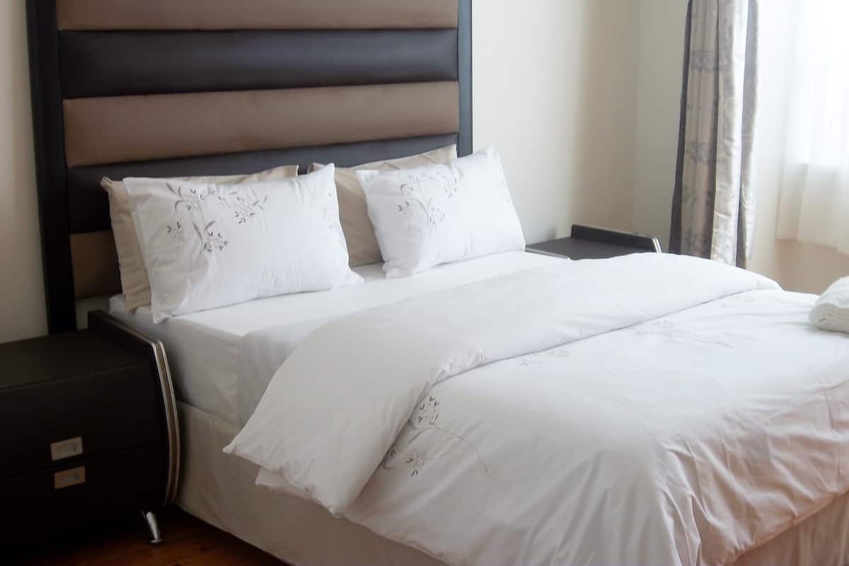 Una cama bien hecha con ropa de cama blanca y almohadas decorativas, un cabecero oscuro y una mesita de noche pequeña con un cajón.