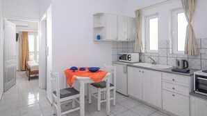 A compact kitchen with white cabinetry, a microwave, a kettle, and a sink. There is a dining area with a table and chairs, and a bed is visible in the adjacent room. - Litsa Mare apartments (Malevizi)