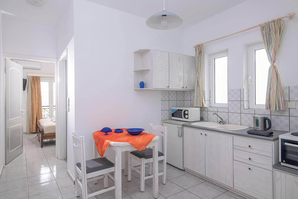 A compact kitchen with white cabinetry, a microwave, a kettle, and a sink. There is a dining area with a table and chairs, and a bed is visible in the adjacent room.