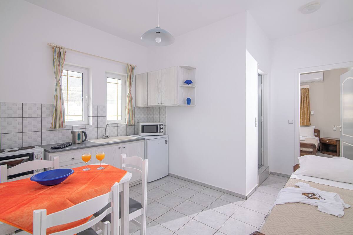 A compact kitchen with a microwave, kettle, and a small refrigerator. There is a dining table with a bowl and two glasses of orange juice.