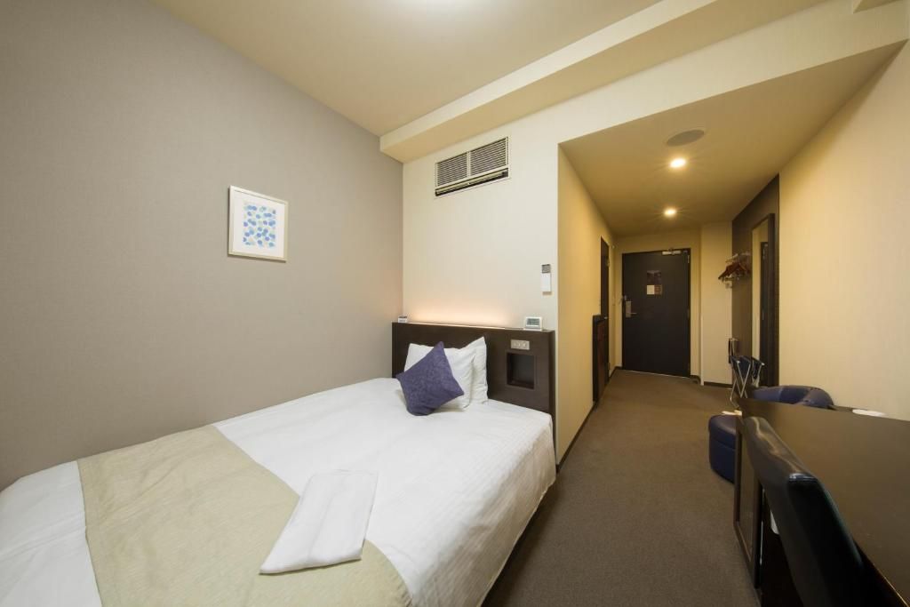 A hotel room with a single bed, a desk, and a chair. There is a hallway leading to a door.