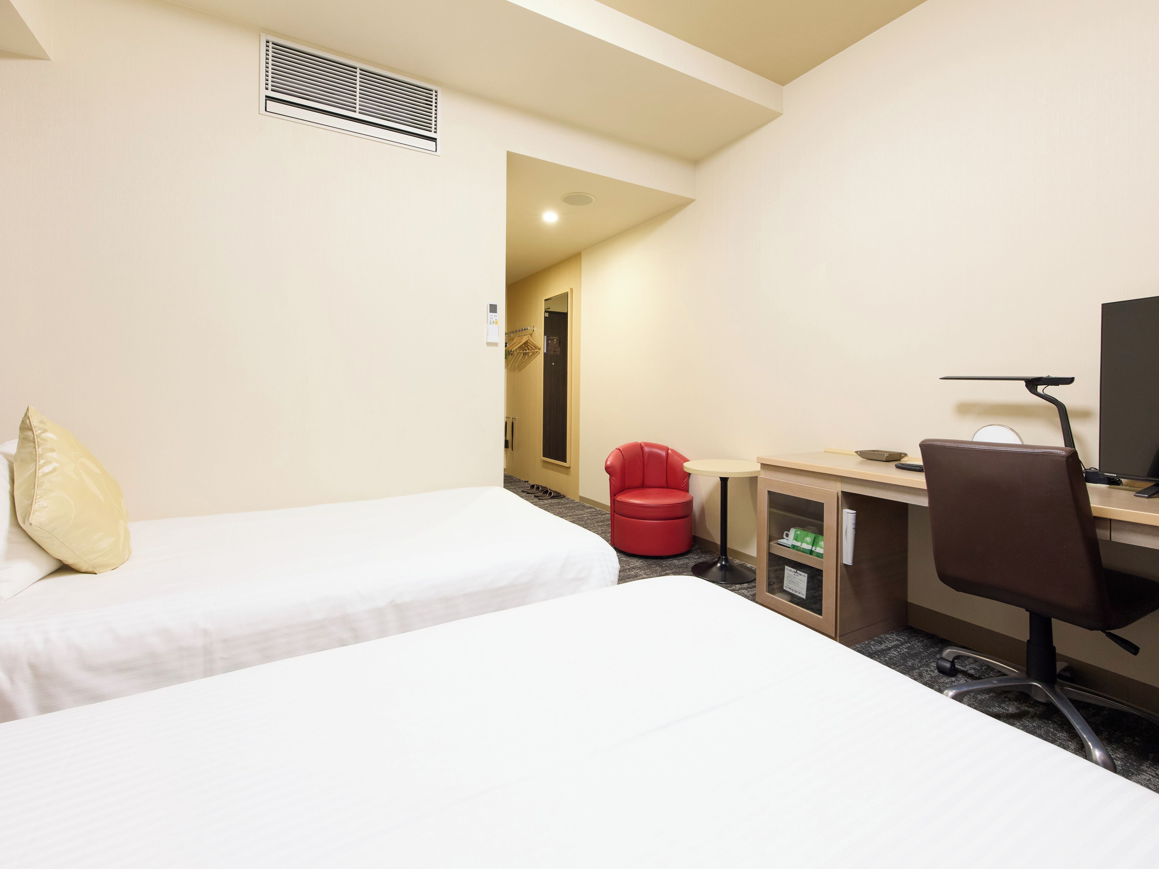 A hotel room with two beds, a desk with a chair, a red chair, a computer, and a TV.
