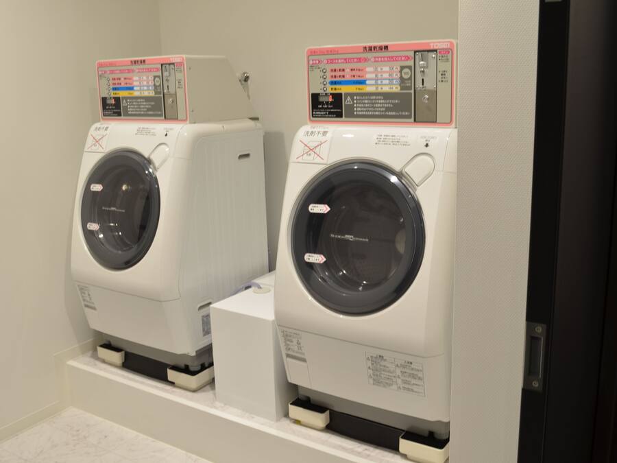 Two front-loading washing machines with control panels, placed side by side in a room with white walls and a tiled floor.